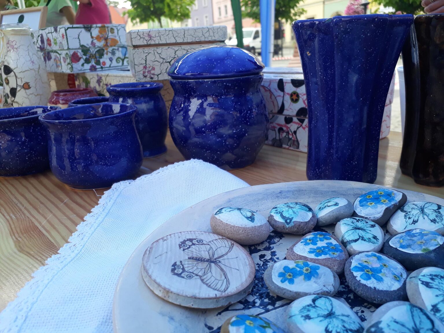 Stoisko plenerowe na którym pokazane są wyroby ceramiczne: niebieskie ceramiczne wazony, donice, kamienie ozdobione technika decoupage w motyle i niezapominajki.