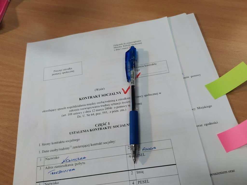 Dokument lezy na biurku. Dokument ma napis kontrakt socjalny. Na dokumencie położony jest długopis w kolorze niebieskim.
