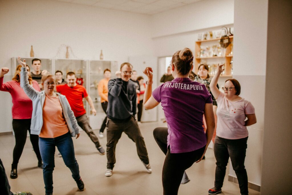 Grupa wykonuje ćwiczenia z fizjoterapeutką. Widać u nich radość. Wykonują ćwiczenia tak jakby tańczyli.