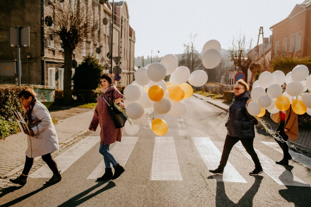 Na zdjęciu przestawione są panie przechodzące przez ulicę. panie trzymają w rękach balony. balony unoszą się nad głowami pań. Panię są radosne i uśmiechnięte. Na drugim planie zdjęcia widać kamienice, które znajdują się na ulicy.