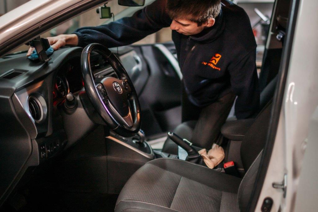 Pokazany środek przedniej części samochodu osobowego, deska rozdzielcza, którą mężczyzna czyści szmatka. Mężczyzna poopiera się o siedzenie w samochodzie.