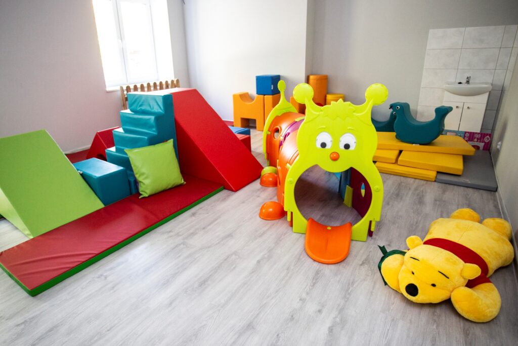 W pomieszczeniu stoją na podłodze zabawki. Zabawki są duże. mają kolory czerwony, żółty. Na podłodze leży pluszowy duży miś. Z tyłu leżą na podłodze materace do zabawy.